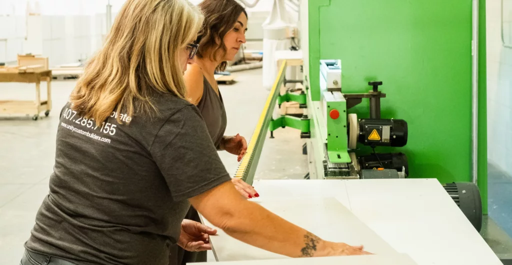 Two cabinetmakers measuring a white panel beside a green edgebander